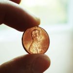 Image of a penny held between two fingers with a white indoor background. Dividend-Paying Penny Stocks